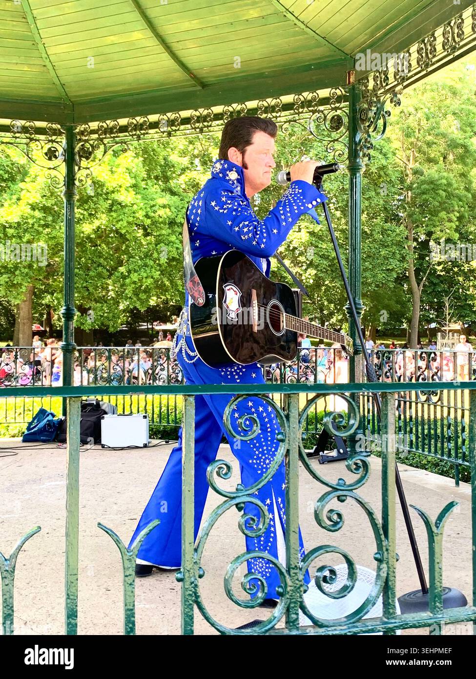 L'artista tributo a Elvis intrattiene la folla all'Abington Park Northampton, Inghilterra, cantando in estate con la folla che guarda vicino alla tribuna - Immagine stock catturata con smartphone
