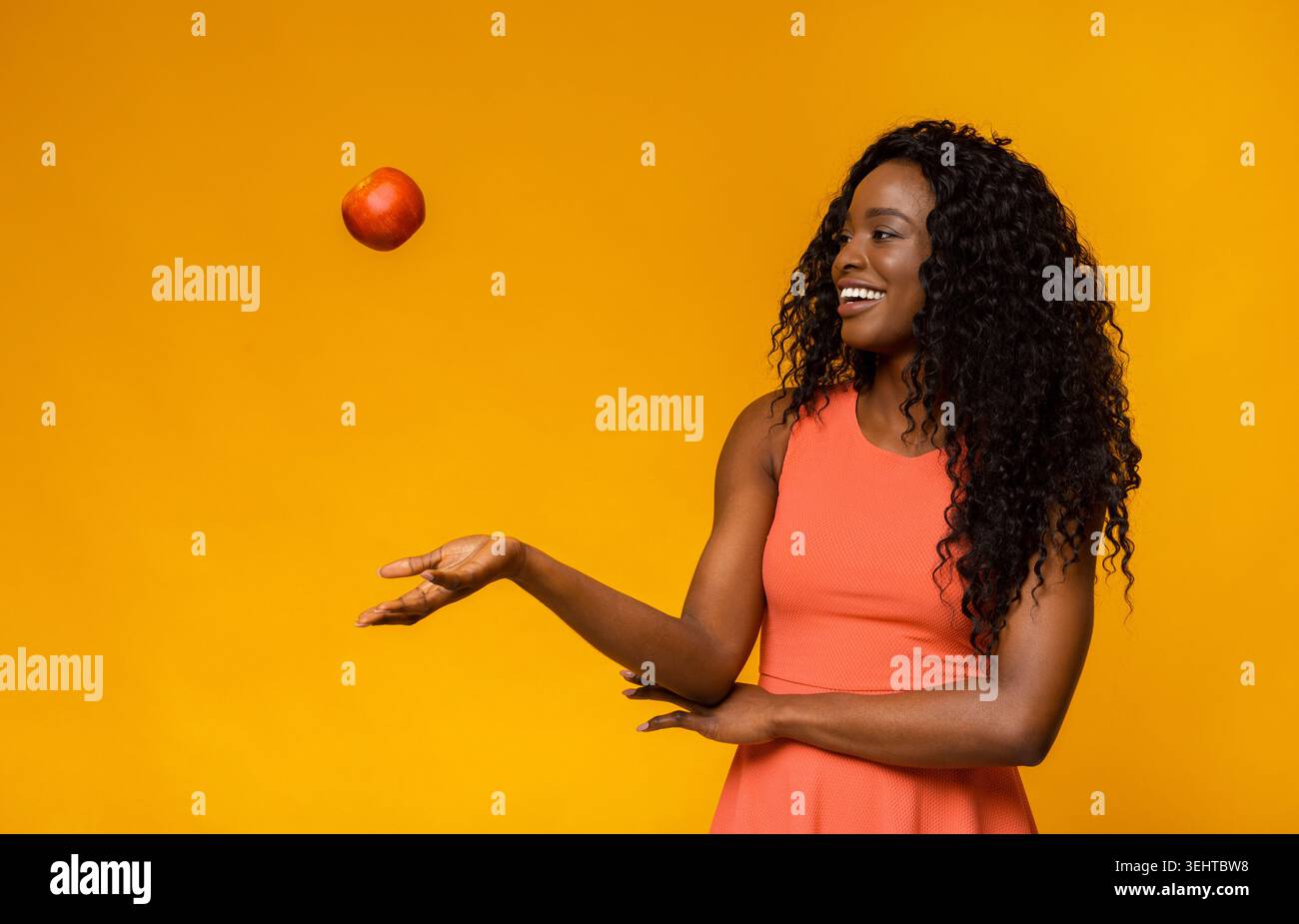 Donna in abito rosa bilancia la mela rossa con un vivace sfondo giallo in un momento di gioco Foto Stock