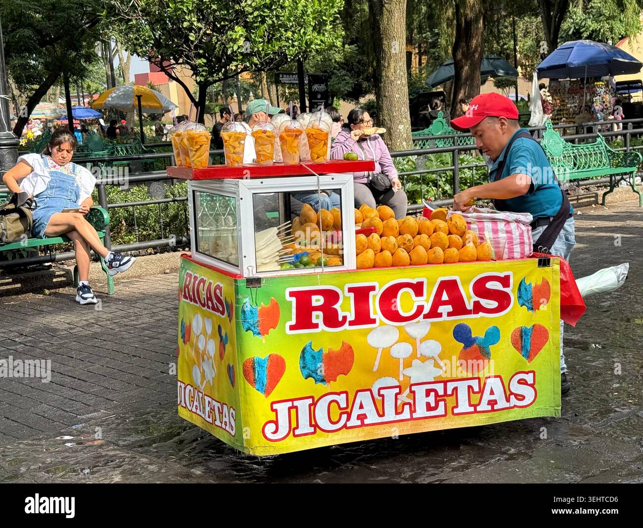 Un venditore ambulante prepara jicaletas e spuntini di frutta da un carrello colorato in una piazza pubblica di Coyoacán, città del Messico, Messico - Immagine stock catturata con smartphone