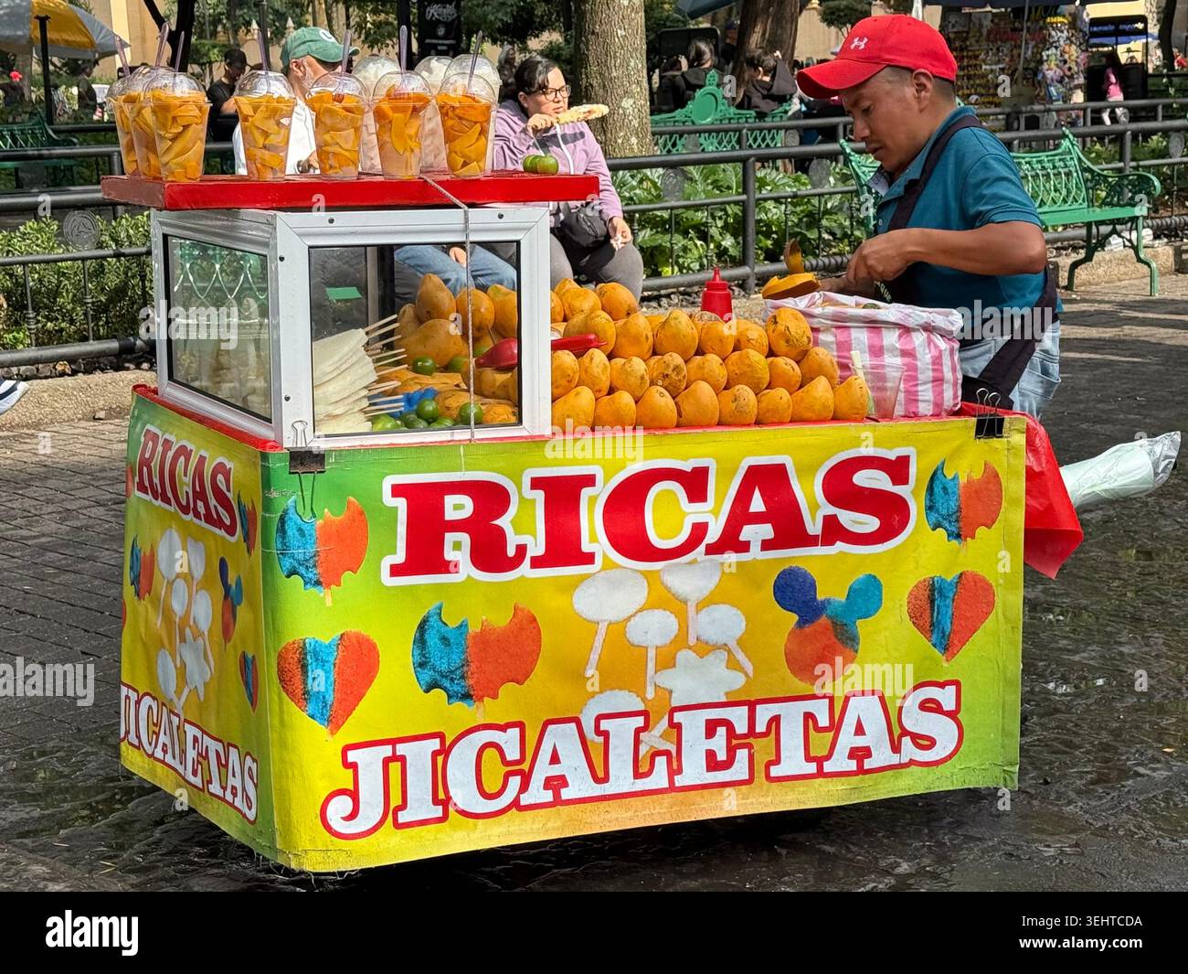 Un venditore ambulante prepara jicaletas e spuntini di frutta da un carrello colorato in una piazza pubblica di Coyoacán, città del Messico, Messico - Immagine stock catturata con smartphone
