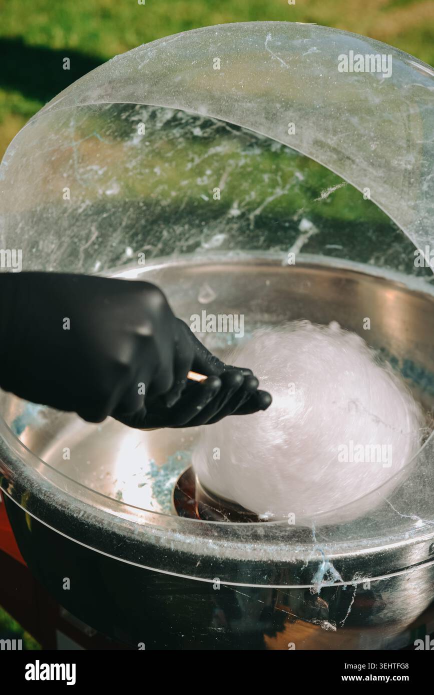 Primo piano di una macchina per caramelle di cotone che filtra caramelle bianche fresche in occasione di una fiera o di un festival all'aperto. Vivace scena gastronomica con guanto a mano Foto Stock