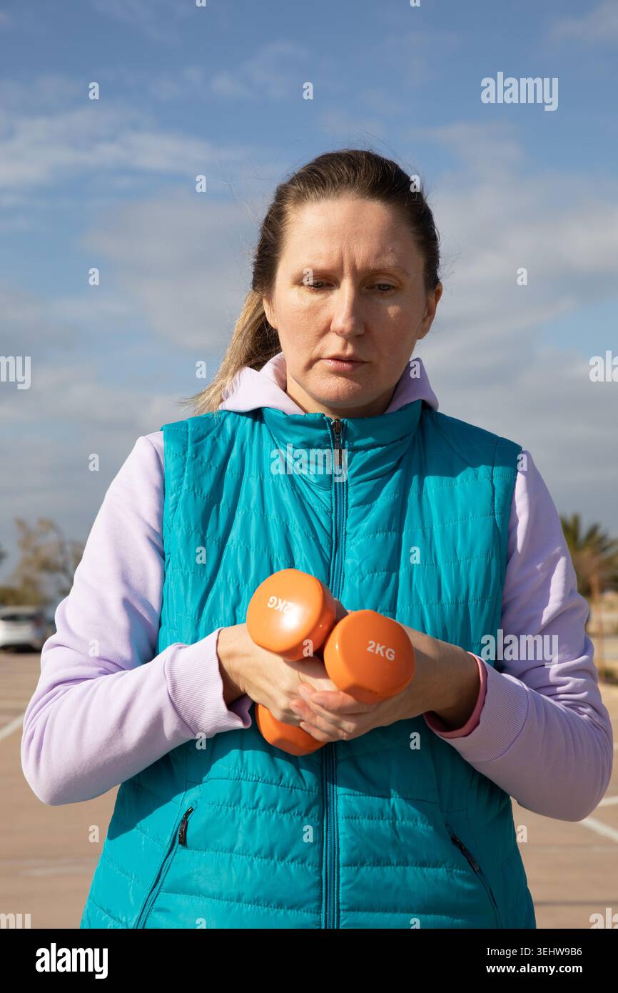 Una donna di mezza età con due manubri arancioni sembra concentrata durante una sessione di allenamento all'aperto nel parcheggio costiero. Il ritratto cattura la concentrazione durante gli allenamenti di fitness nelle giornate di sole all'aperto Foto Stock