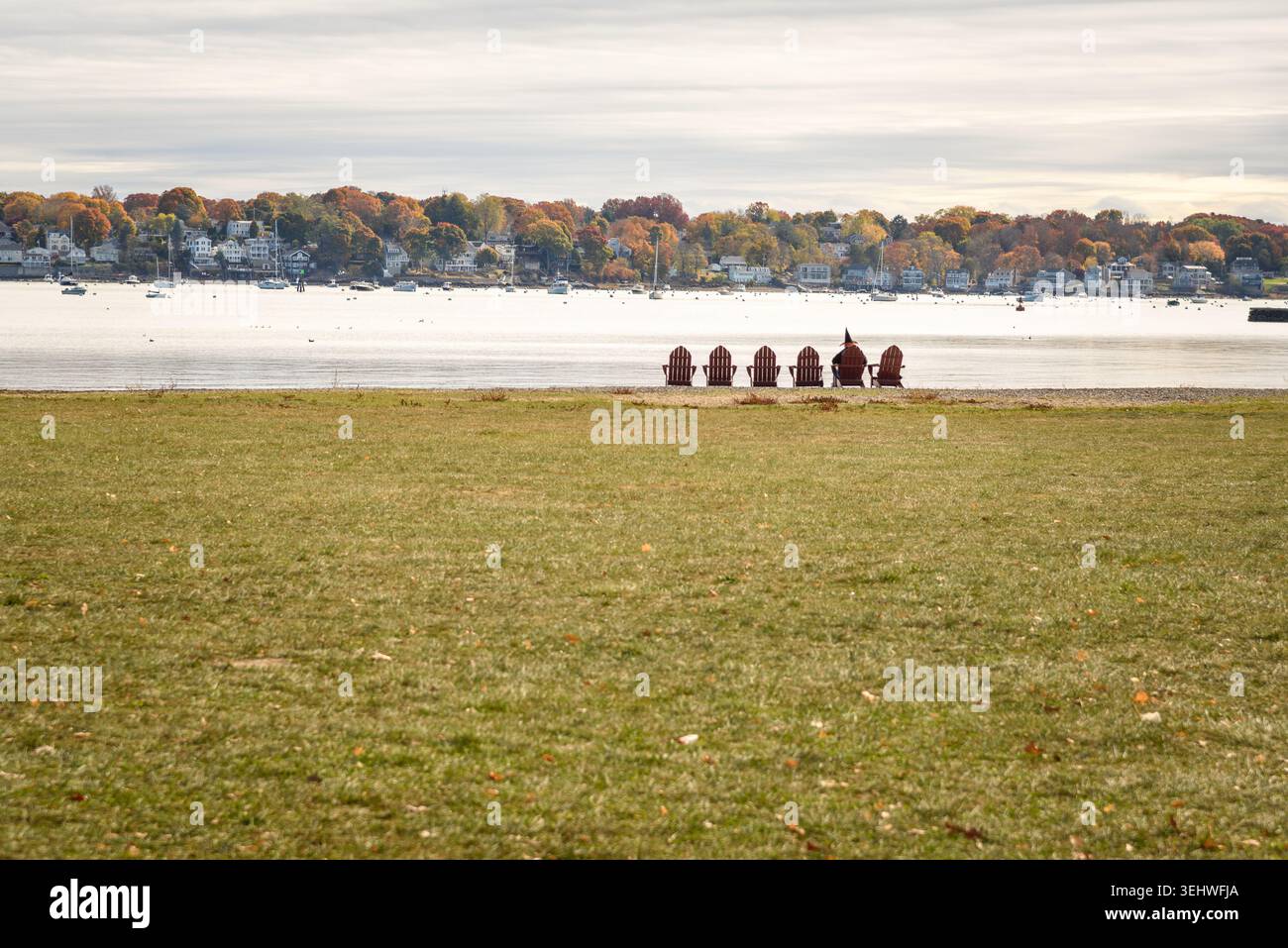 Fila di sedie Adirondck sull'erba di fronte a un porto naturale in una soleggiata mattinata d'autunno. Una persona vestita da strega è seduta su una delle sedie. Foto Stock