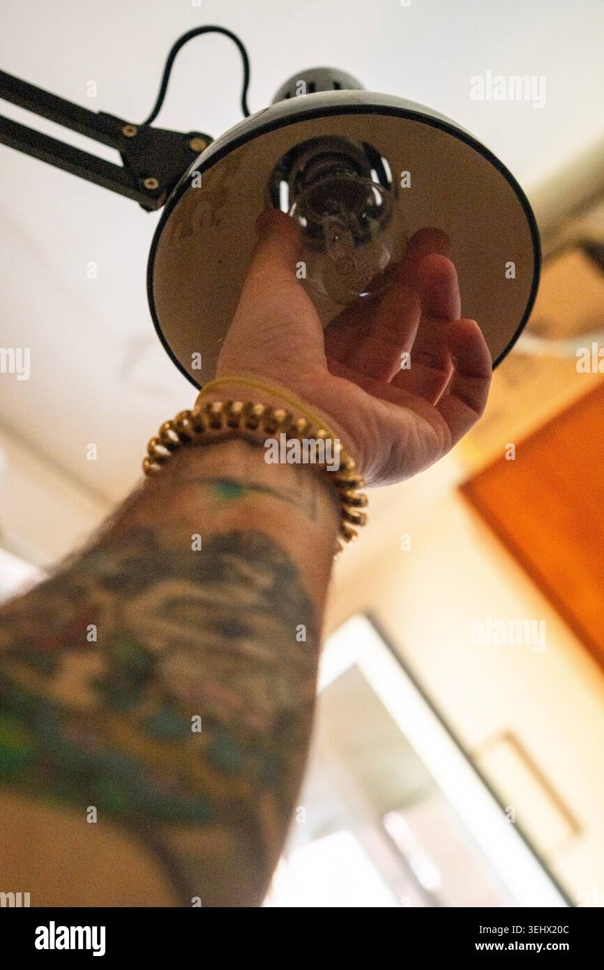 La mano di un uomo incassa la lampadina di una lampada da scrivania. L'avambraccio dell'uomo è tatuato. L'immagine richiama diversi concetti, tra cui "fai da te" e "elettricit" Foto Stock