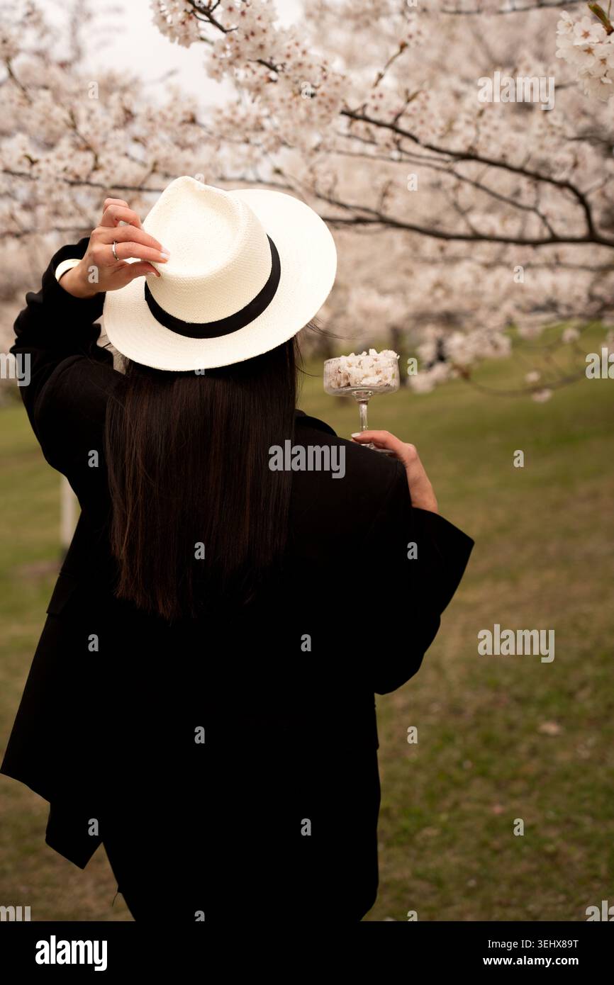 vista posteriore di una persona con un cappotto nero che regola un cappello bianco a teglia larga mentre tiene in mano un bicchiere coupé riempito di petali di ciliegio sotto soft pi Foto Stock