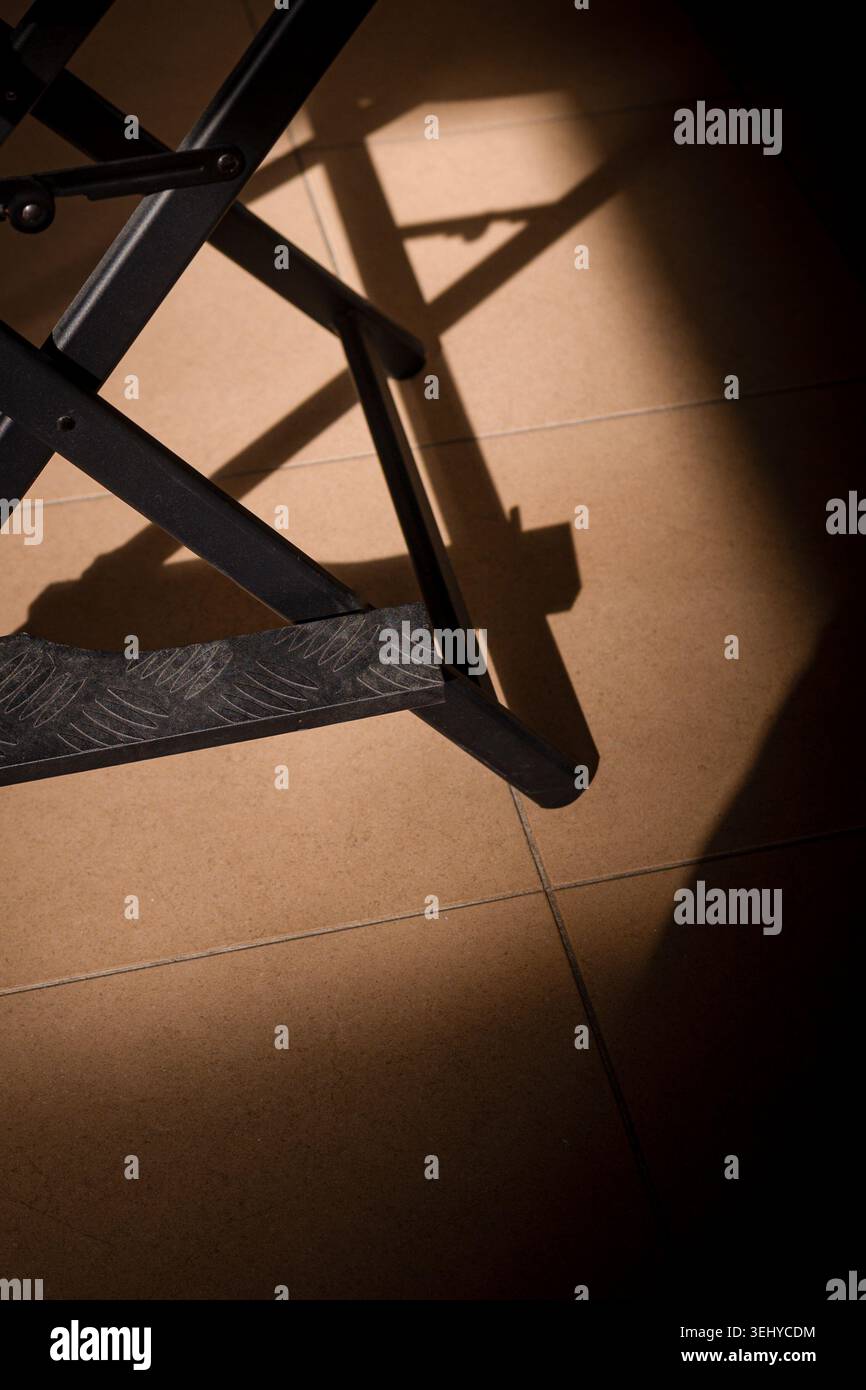 Struttura in metallo e poggiapiedi modellato di una sedia pieghevole che crea forti forme geometriche e ombre su piastrelle di colore marrone chiaro Foto Stock