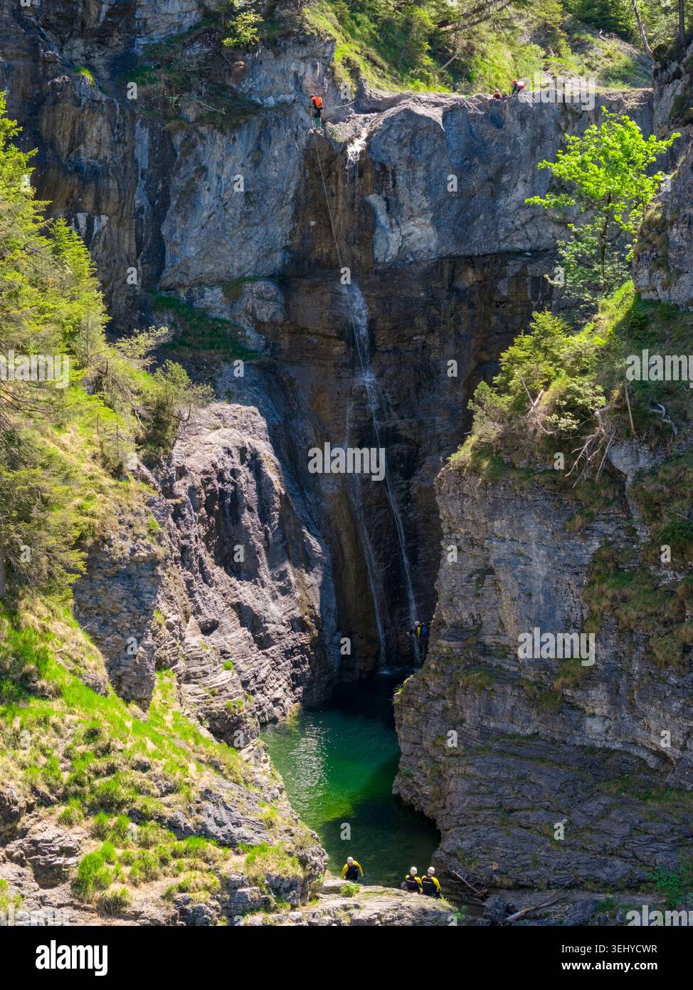 Un'esperta squadra di canyoning si arrampica lungo una ripida gola rocciosa in una piscina color smeraldo sotto una stretta cascata, mostrando sport d'avventura, lavoro di squadra, sa Foto Stock