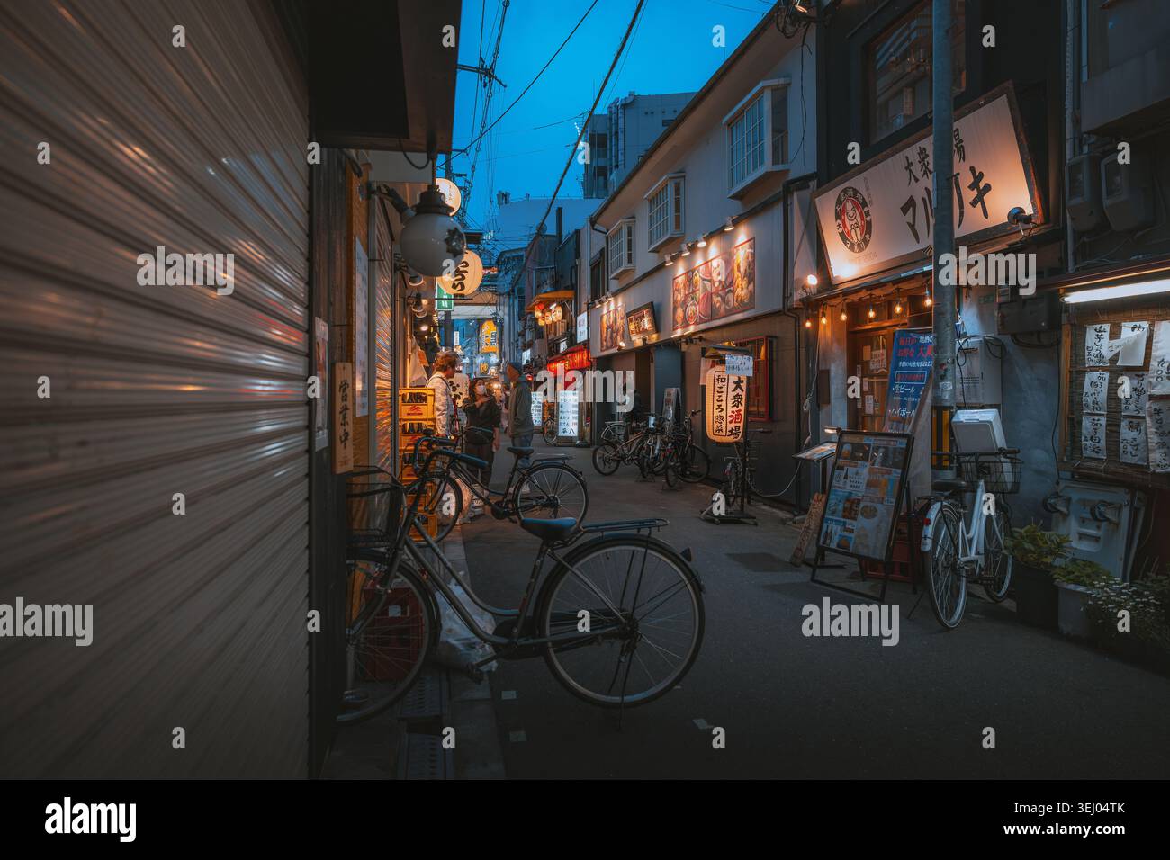 Osaka, Giappone - 26 aprile 2026: Vista di uno stretto vicolo al crepuscolo con cartelli illuminati, biciclette parcheggiate e persone che camminano per la strada. Foto Stock
