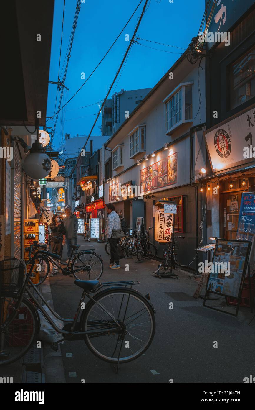 Osaka, Giappone - 26 aprile 2026: Vista di uno stretto vicolo al crepuscolo con lanterne tradizionali, biciclette parcheggiate e persone che camminano accanto a negozi locali e ristoranti Foto Stock