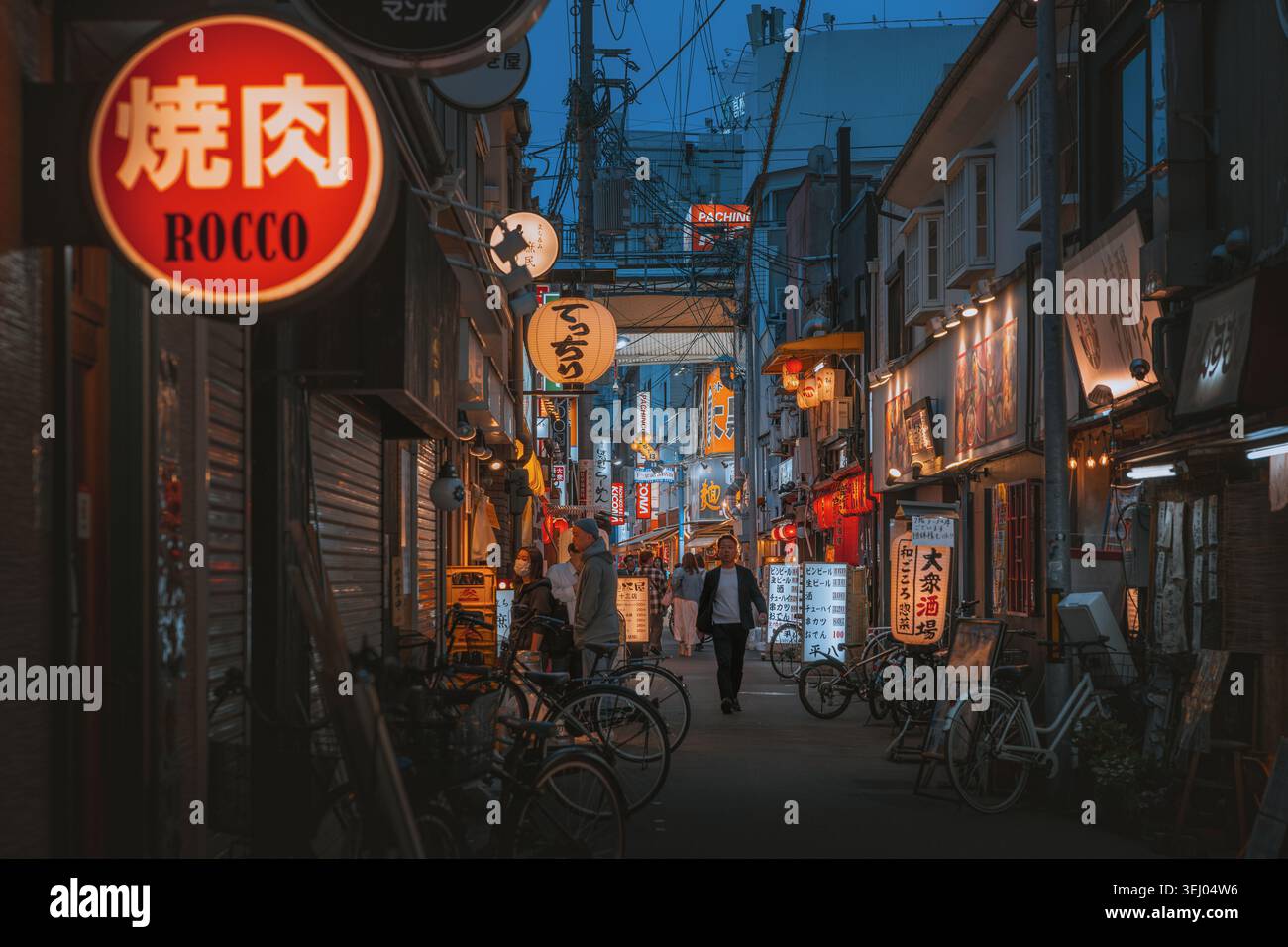 Osaka, Giappone - 26 aprile 2026: Vista di uno stretto vicolo al crepuscolo con insegne al neon brillanti, lanterne tradizionali e persone che camminano accanto alle biciclette parcheggiate Foto Stock