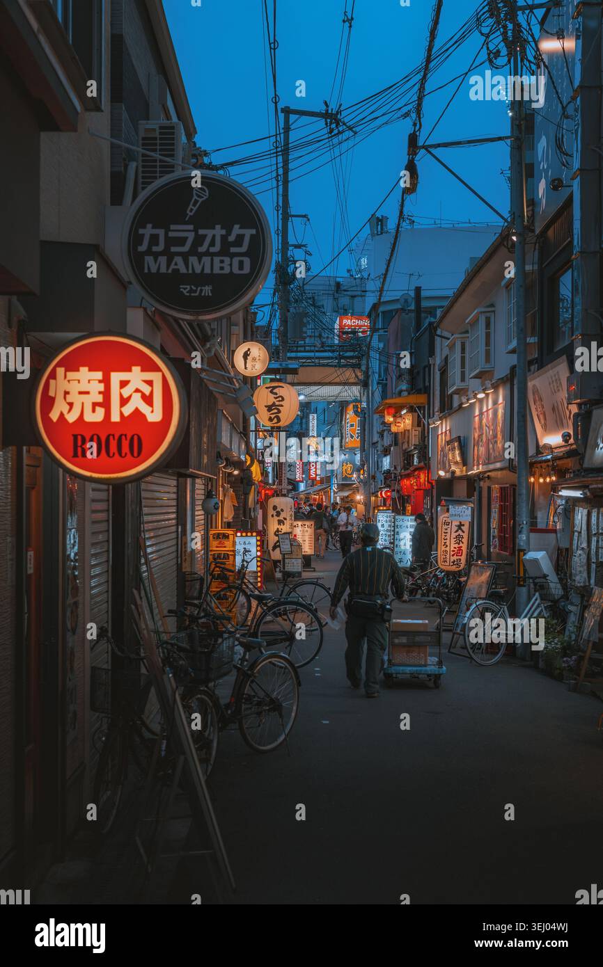 Osaka, Giappone - 26 aprile 2026: Vista di un vicolo stretto al crepuscolo con cartelli luminosi per ristoranti, biciclette parcheggiate e un uomo che spinge un carrello per le consegne Foto Stock