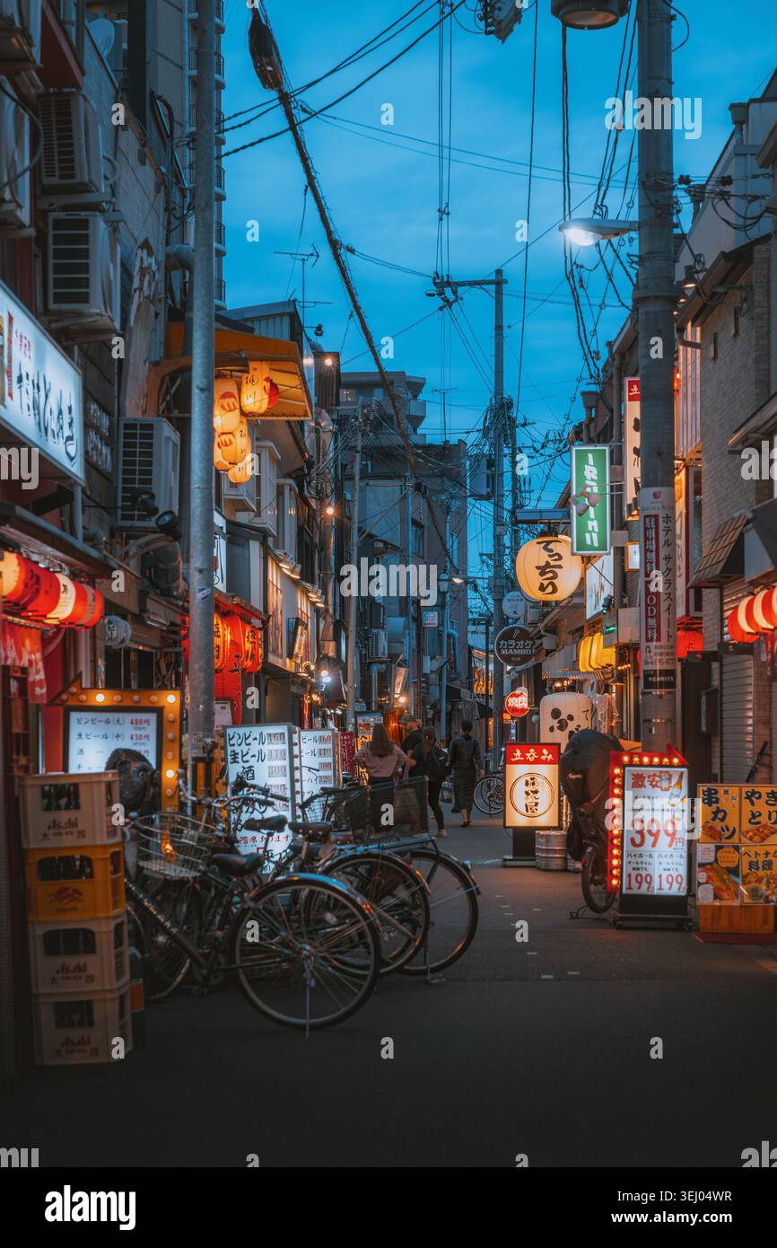 Osaka, Giappone - 26 aprile 2026: Veduta di una stradina stretta al crepuscolo con lanterne luminose, insegne tradizionali giapponesi, biciclette parcheggiate e persone che camminano a piedi Foto Stock