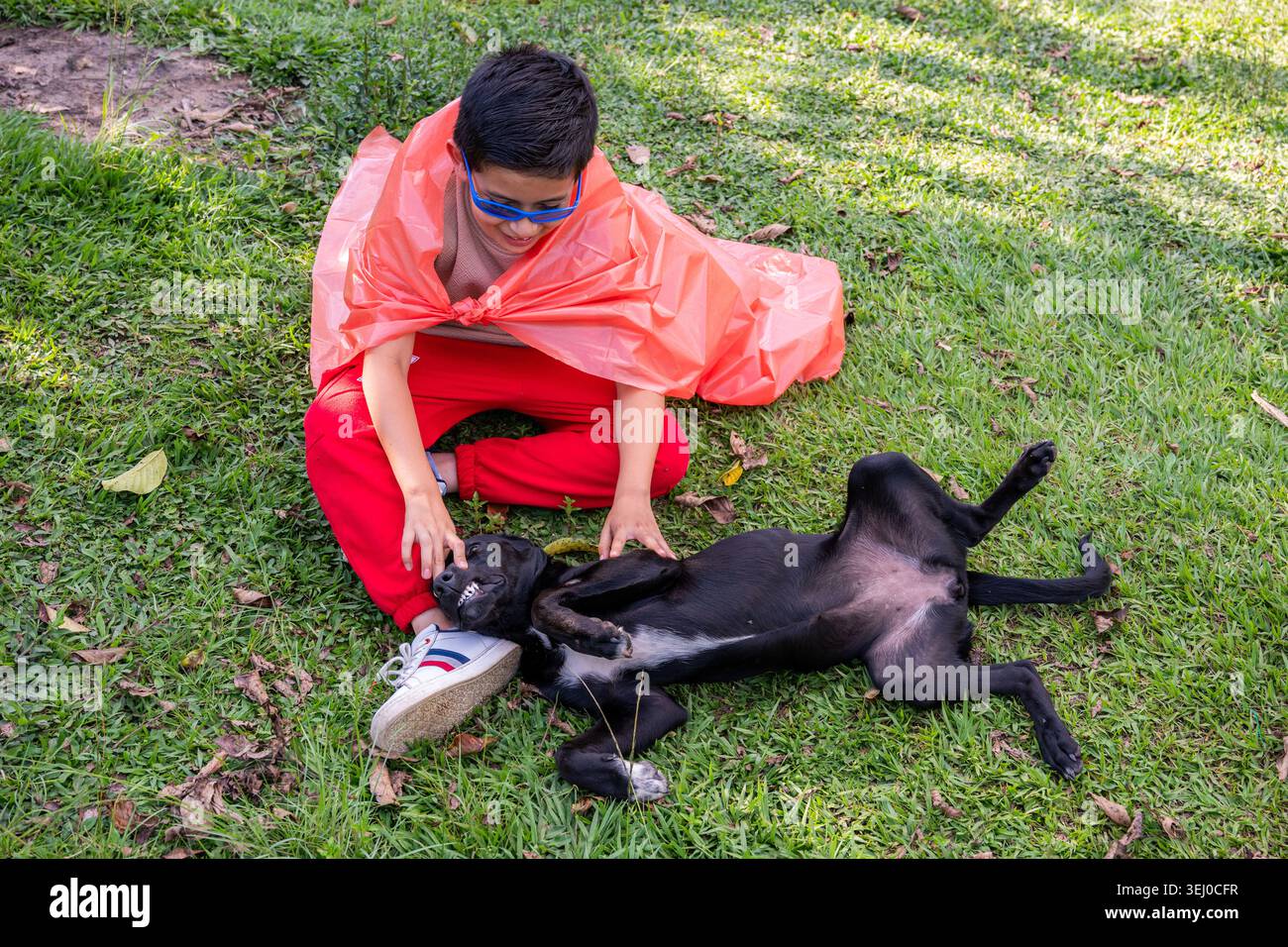 Ragazzo con mantello arancione e occhiali blu che ride con un cane nero felice che rotola giocosamente sull'erba verde del parco, divertimento estivo spensierato e fantasioso youthfu Foto Stock