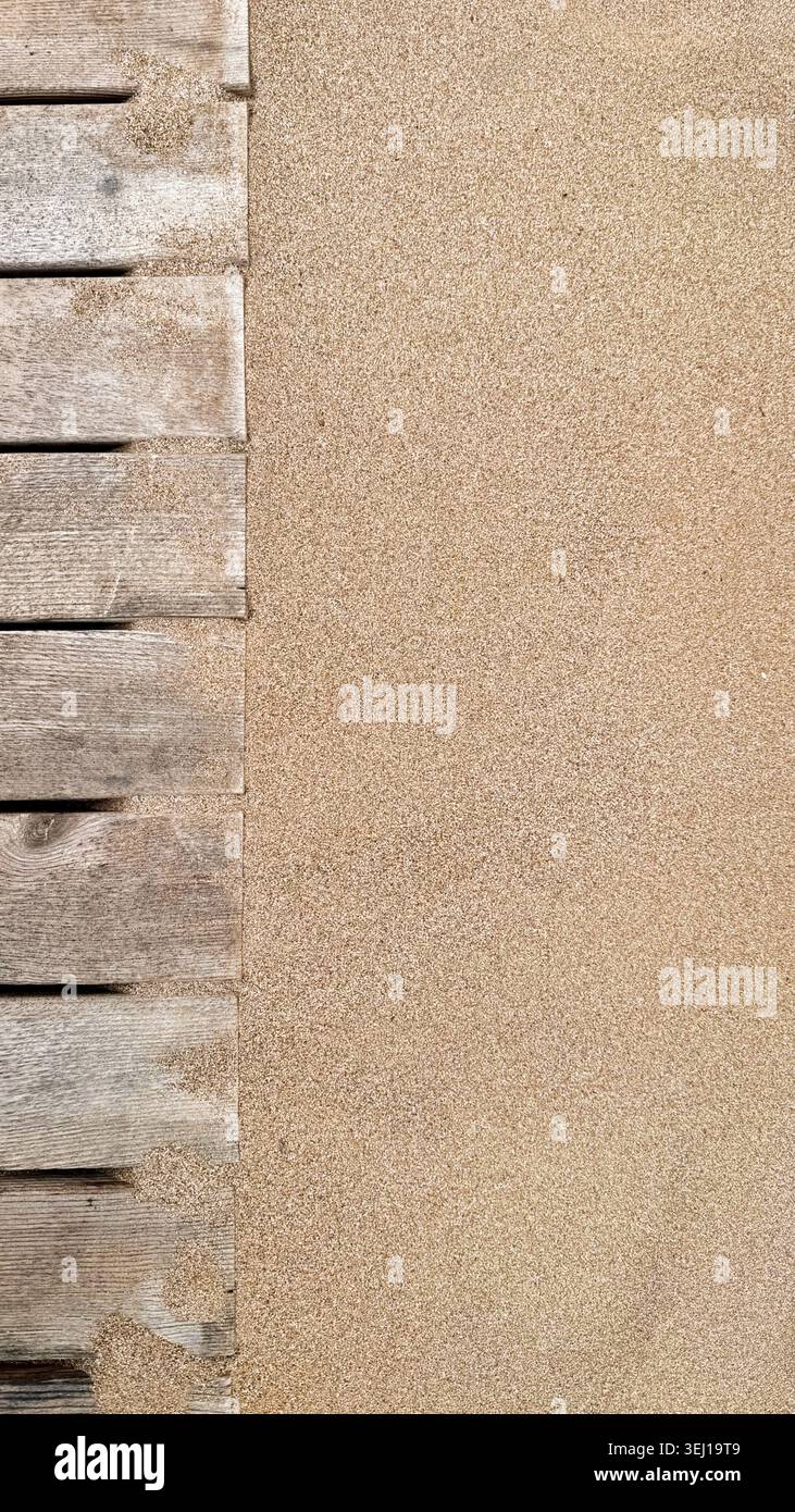 Superficie sabbiosa ruvida adiacente a tavole di legno intemperie, che mostra elementi naturali in un ambiente costiero o spiaggia con una tavolozza di colori caldi Foto Stock