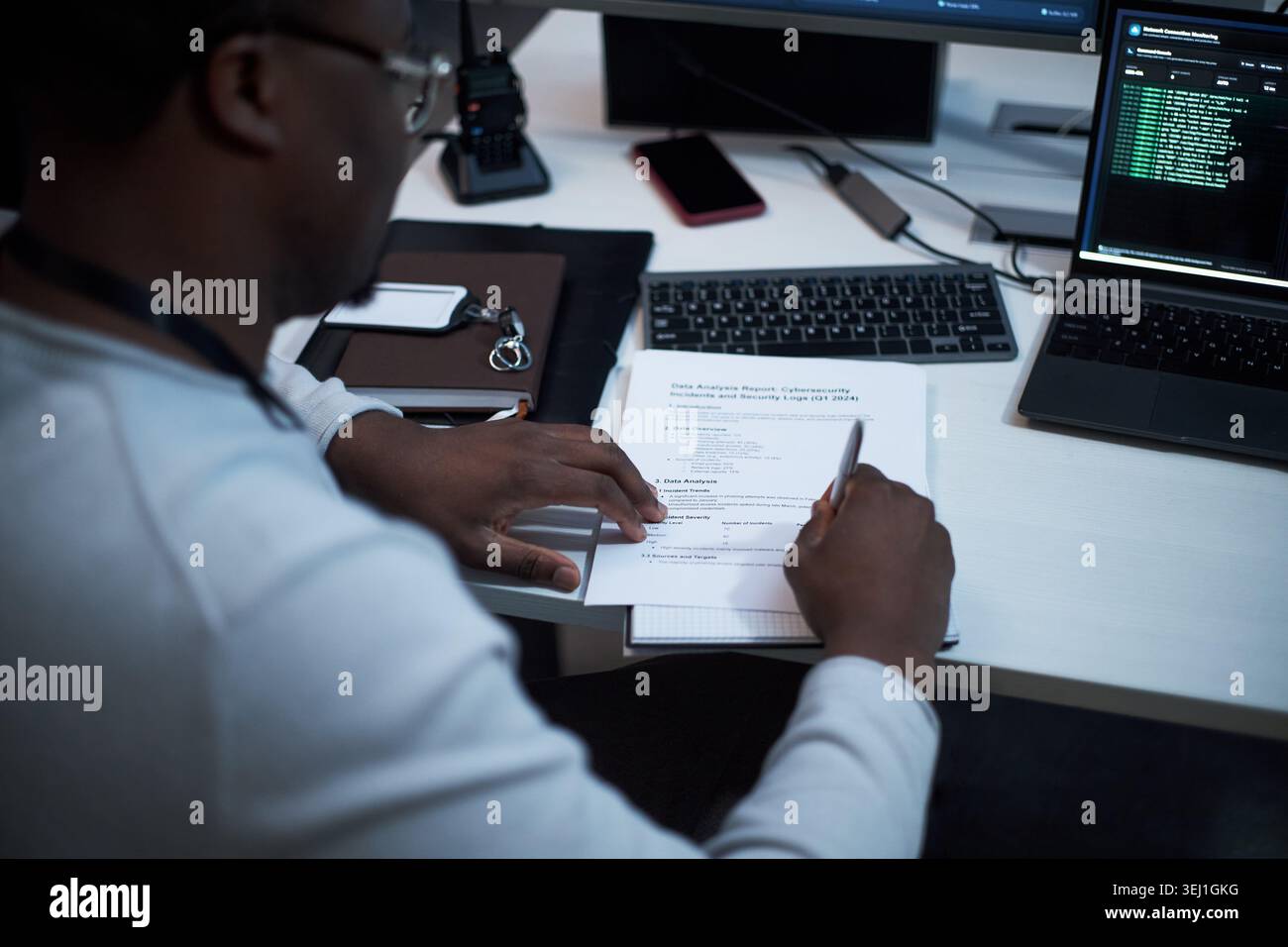 Foto alla spalla di un giovane adulto nero che esamina il rapporto sulla sicurezza informatica e lo schermo di codifica, prendendo appunti alla postazione di lavoro. Adatto per la sicurezza IT, la verifica dei rischi e il marketing della conformità Foto Stock