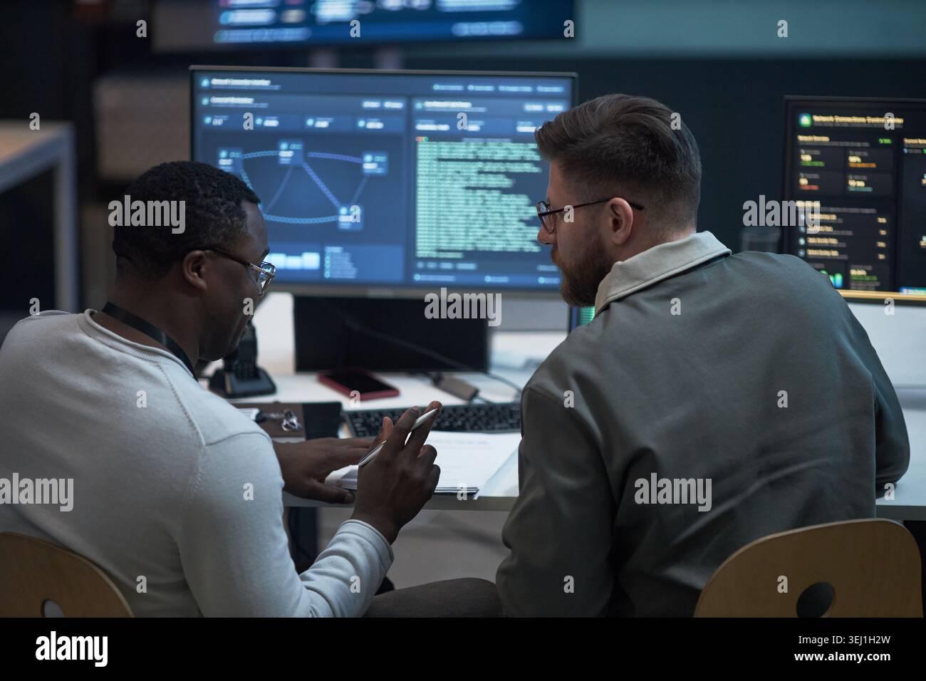 Giovane adulto afroamericano e giovane adulto caucasico che esamina i dati di sicurezza informatica su monitor, collaborando in ufficio. Per le operazioni IT, la sicurezza della rete, il marketing di squadra Foto Stock