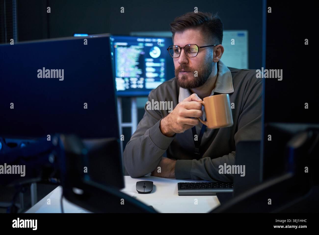 Giovane uomo adulto caucasico che monitora il codice sugli schermi del desktop mentre beve caffè durante il turno di lavoro tardivo, rappresentando il lavoro di sicurezza informatica. Ideale per personale IT, servizi software, supporto tecnico Foto Stock