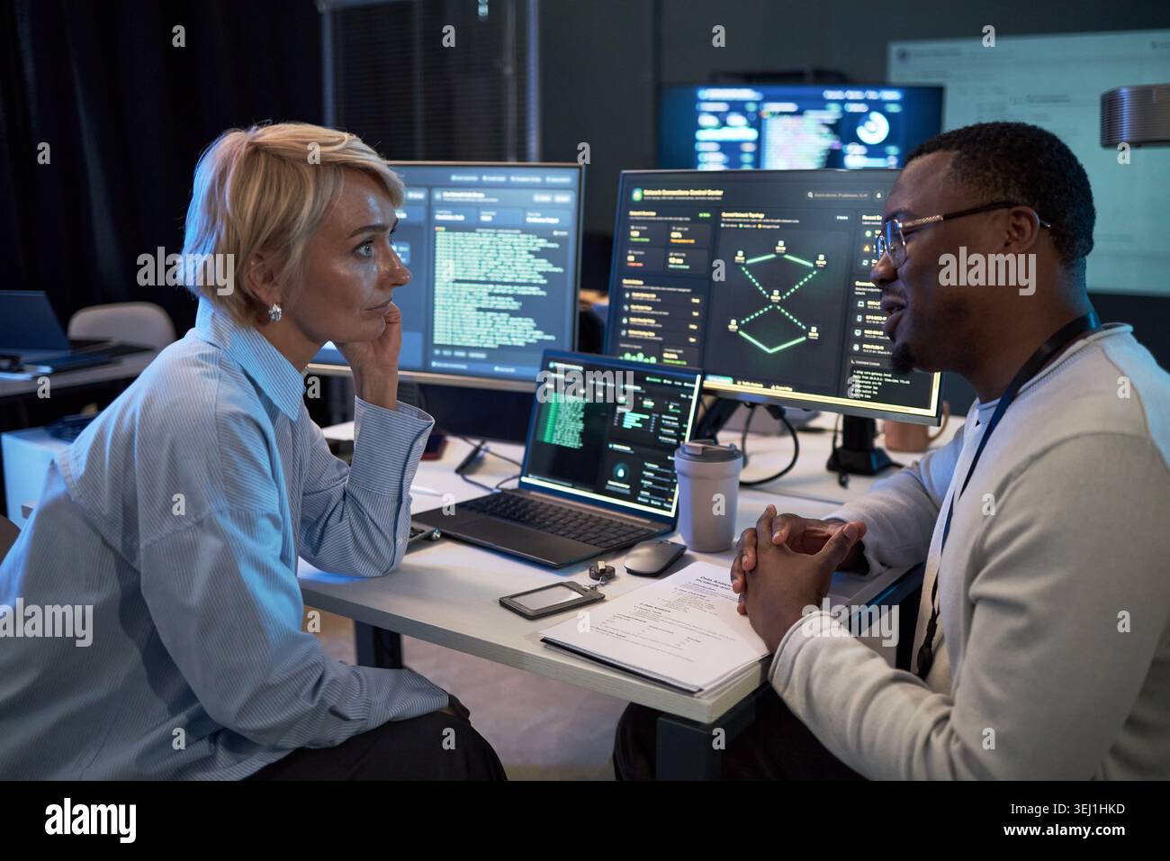 Donna caucasica di mezza età e uomo nero discutono dei dati sulla sicurezza informatica presso la workstation, esaminando il codice e i dashboard di rete. Utile per il lavoro di squadra IT, la difesa informatica, le operazioni tecnologiche Foto Stock