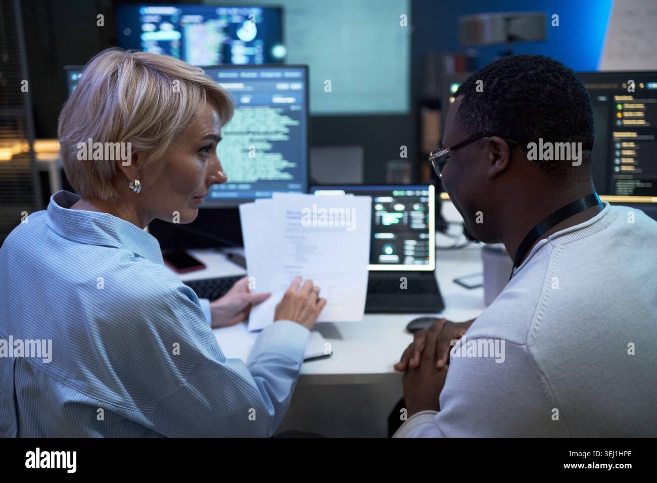 Donna caucasica di mezza età e uomo di colore che esaminano documenti e dati informatici presso la workstation, discutendo di analisi della sicurezza informatica. Ideale per il lavoro di squadra IT, la protezione dei dati e la tecnologia aziendale Foto Stock