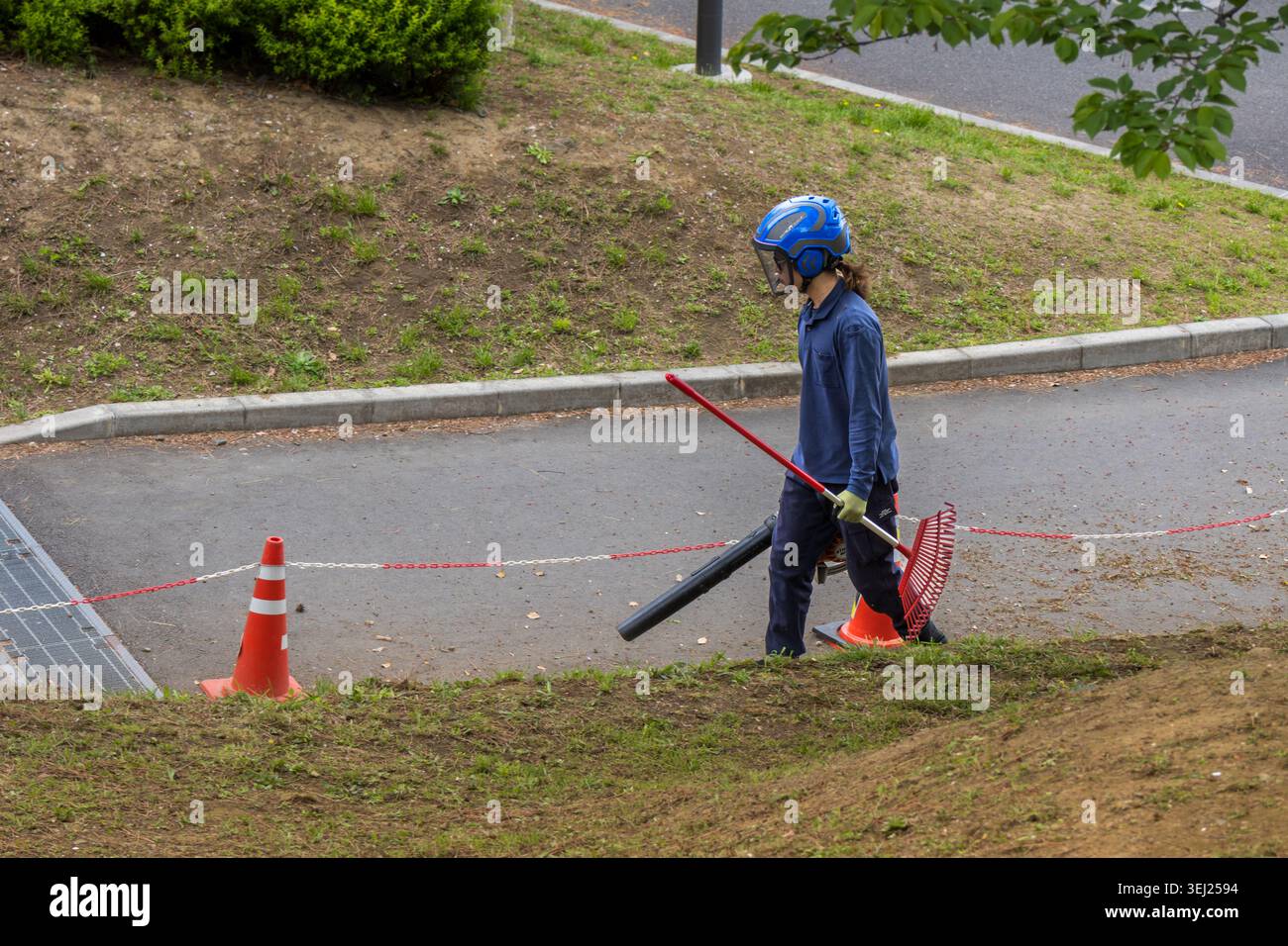Tokyo, Giappone 15 aprile 2026 operatore di manutenzione della città che indossa un casco di sicurezza trasportando un rastrello e un soffiatore di foglie mentre pulisce un percorso pavimentato in un paesaggio paesaggistico Foto Stock