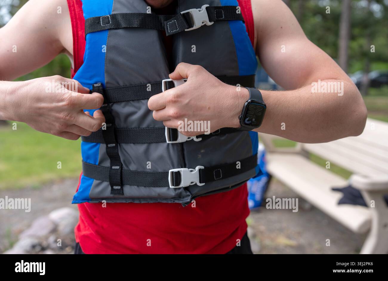 Una fotografia ravvicinata di un uomo in forma che regola e si aggancia un giubbotto salvagente blu e grigio (dispositivo di galleggiamento personale) all'aperto. Indossa una canotta rossa Foto Stock
