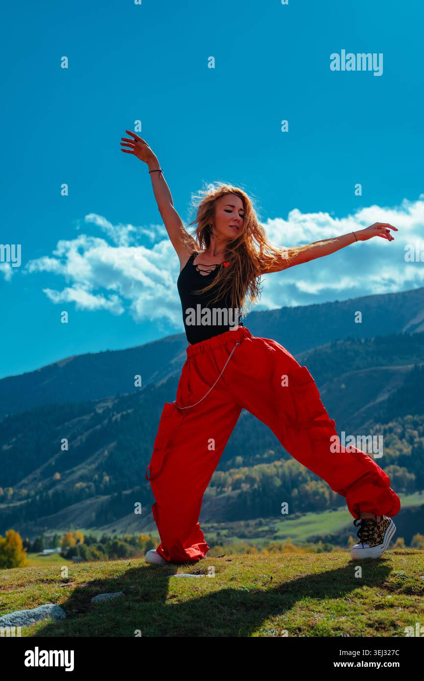 Giovane modella bionda in un abito elegante che si diverte e balla su una collina erbosa con montagne e foreste alle spalle Foto Stock