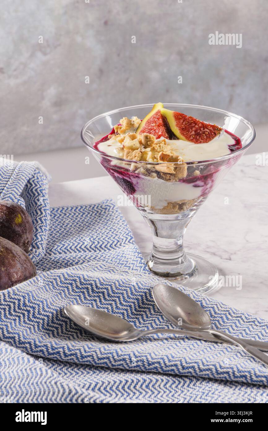 Budino di fichi parfait con yogurt, marmellata di mirtilli, fichi, nocciole e biscotti in vetro su sfondo di marmo bianco Foto Stock
