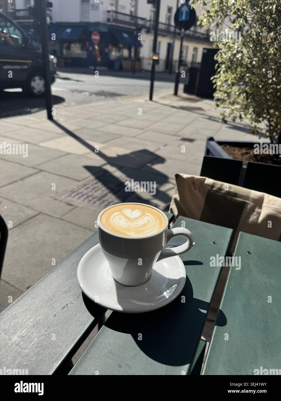 Una tazza bianca di cappuccino caldo in ceramica con un bellissimo motivo a forma di cuore di latte art, seduta su un tavolo in legno verde scuro in un caffè all'aperto a Londra - Immagine stock catturata con smartphone