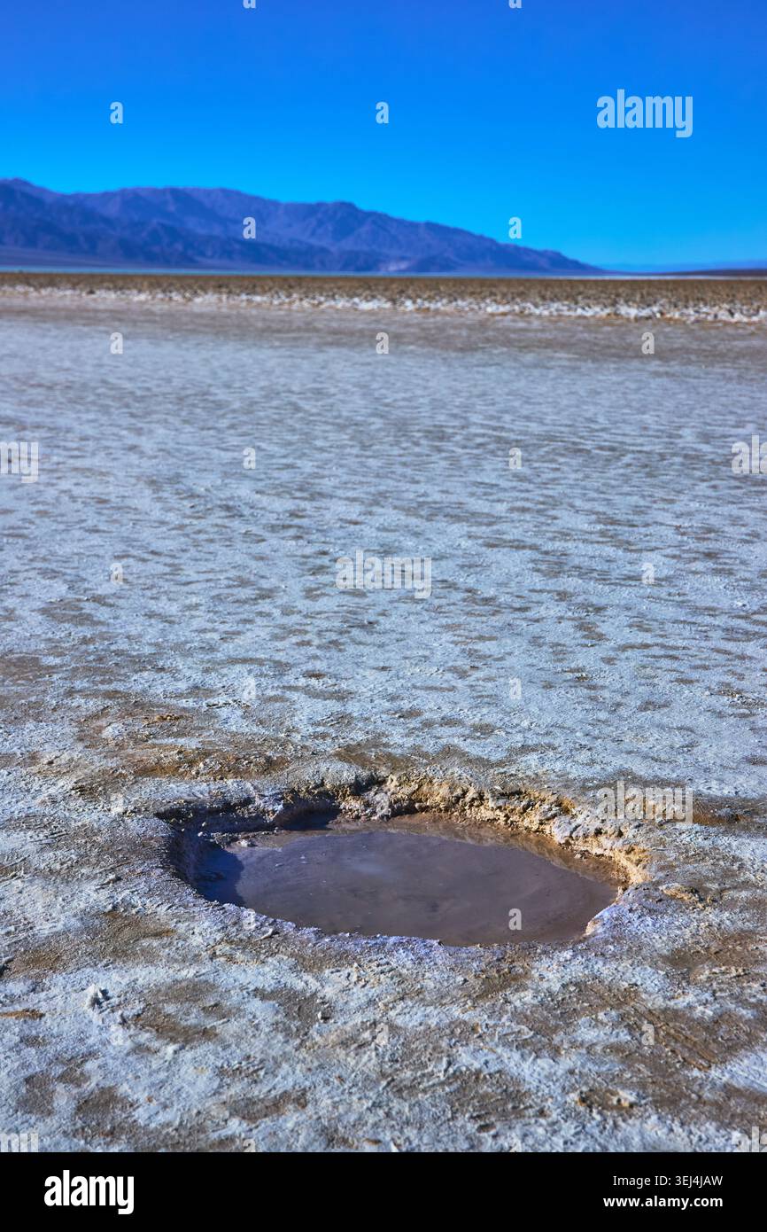 Piscina con acqua salata e bacino di Badwater con montagne lontane sotto il cielo blu Foto Stock