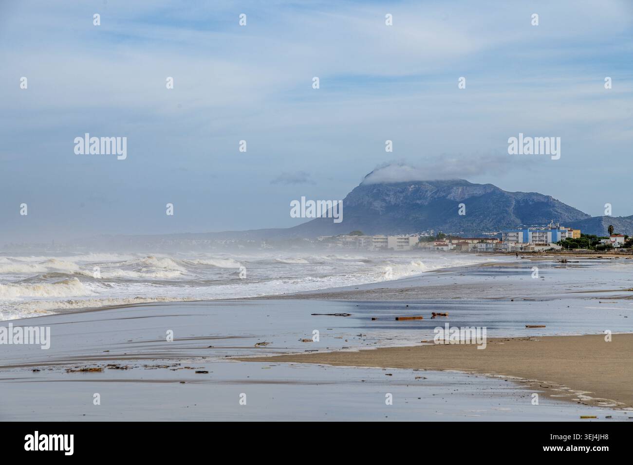 Ampia spiaggia dopo la tempesta, onde che si infrangono, nebbioso sfondo di montagne, edifici costieri, zone sparse, e alghe sulla sabbia bagnata Foto Stock