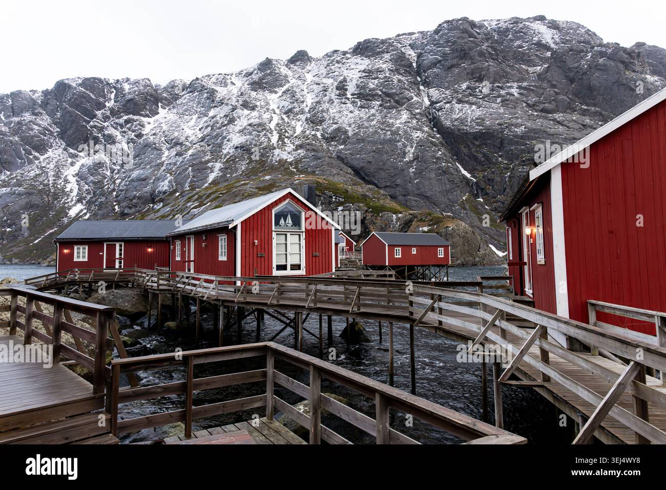 Una vista panoramica invernale delle storiche cabine rosse dei pescatori di rorbu collegate da passerelle in legno sopraelevate e piattaforme a palafitte sull'acqua, il Nusfjord Foto Stock