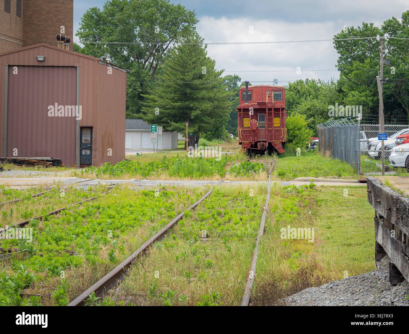 Un vecchio caboose rosso della ferrovia parcheggiato su binari sovradimensionati accanto a un edificio industriale e a una linea di recinzione sotto un cielo nuvoloso Foto Stock