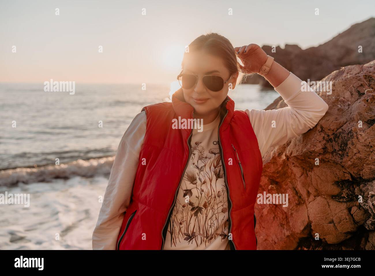 Donna al tramonto, modella femminile in un gilet rosso e occhiali da sole si staglia su una spiaggia rocciosa durante l'ora d'oro sul mare Foto Stock