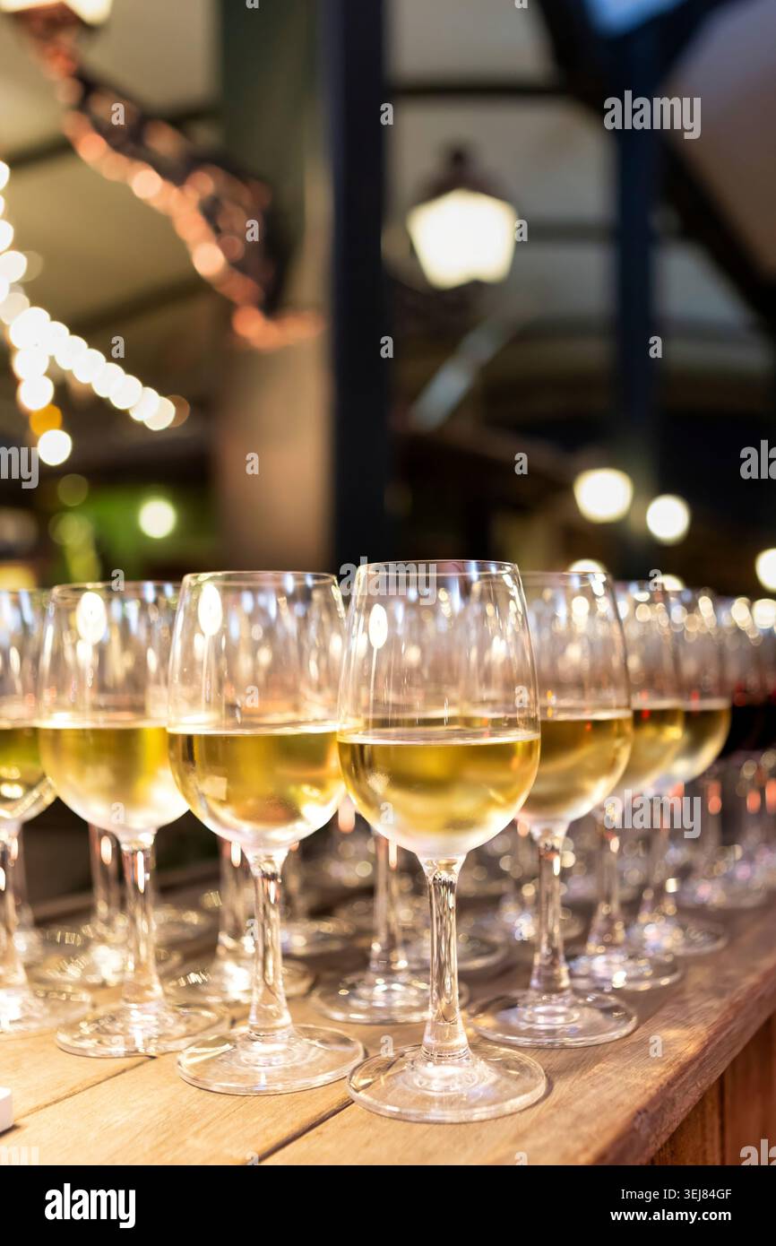 File di bicchieri da vino bianco riempiti allineati su un bancone bar in legno, con calde luci bokeh che si accendono sullo sfondo ad un ricevimento serale. Foto Stock