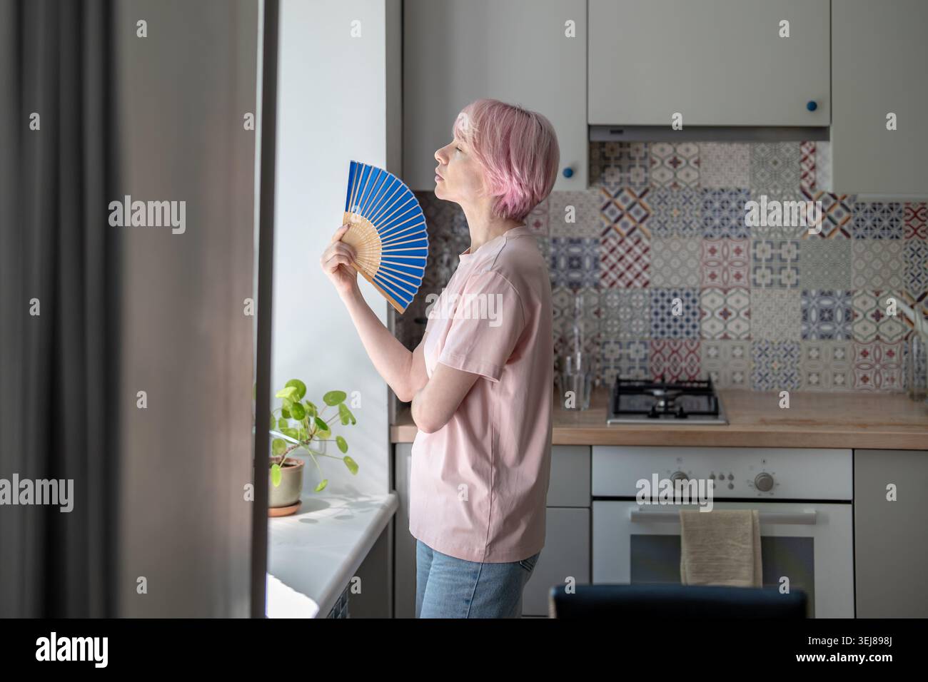 Stanca donna di mezza età in piedi in cucina accanto a una finestra utilizza un ventilatore portatile per alleviare il calore intenso. Onde di calore, aria condizionata non disponibile, soffocamento al coperto Foto Stock
