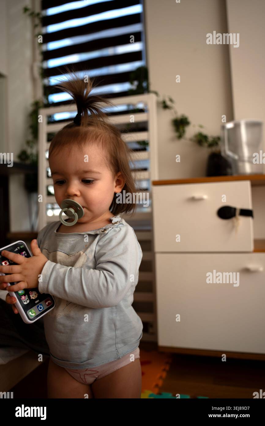 bambino con topknot e succhietto che stringe uno smartphone giocattolo in un accogliente interno illuminato dal sole, tonalità calde neutre, persiane a righe e piante in vaso in t Foto Stock