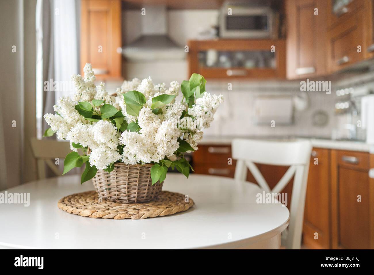 Fresco bouquet di lilla bianco in cesto di vimini sul tavolo da cucina rotondo con interni accoglienti e moderni, luce naturale, arredamento primaverile, stile di vita rustico Foto Stock