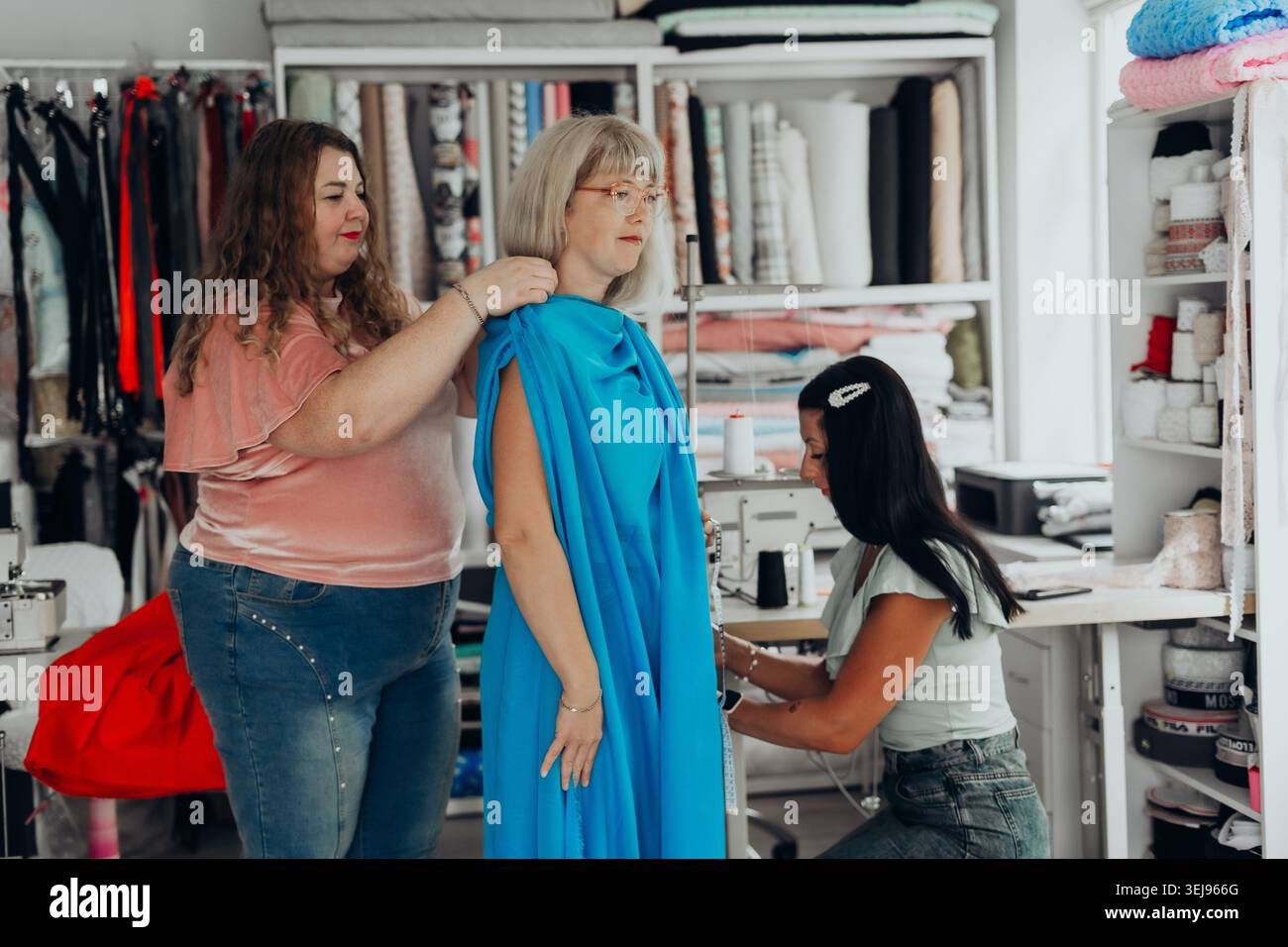 Gli stilisti collaborano in uno studio creativo, indossando un abito blu brillante su una modella donna matura. Sartoria, lavoro di squadra e tendenze di stile moderno Foto Stock