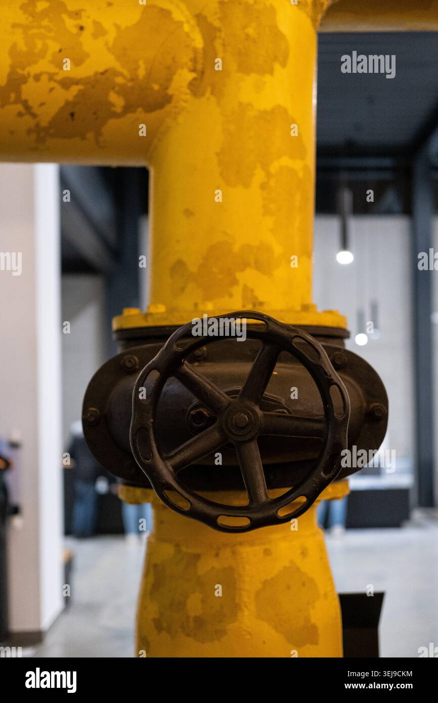 La rotella della valvola in metallo nero è montata su un tubo industriale giallo presso uno stabilimento di macchinari per interni. Foto Stock