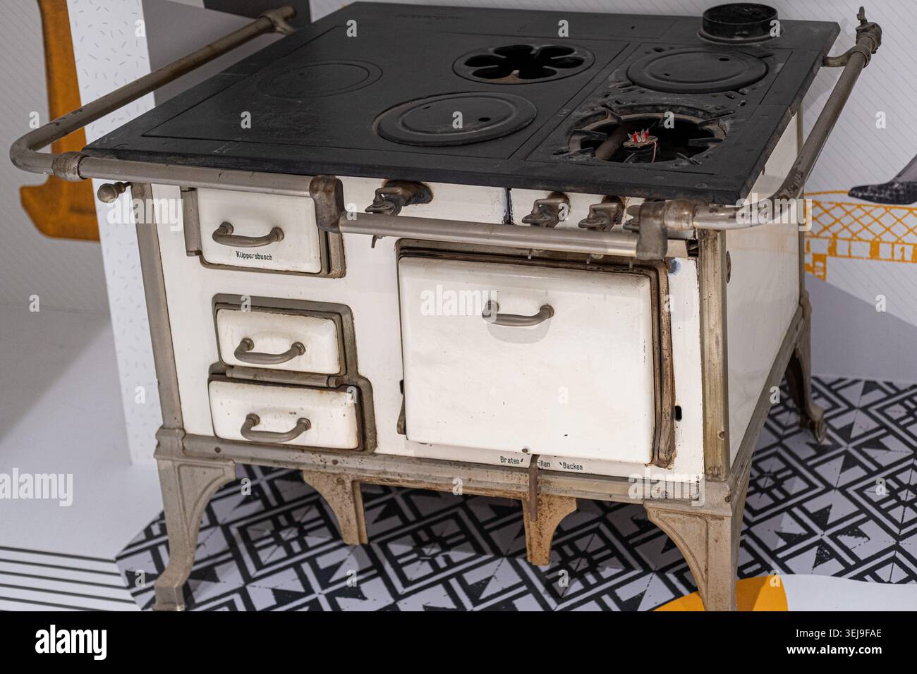 Una stufa vintage a smalto bianco con una superficie di cottura nera è esposta su un pavimento a motivi geometrici in un museo interno. Foto Stock