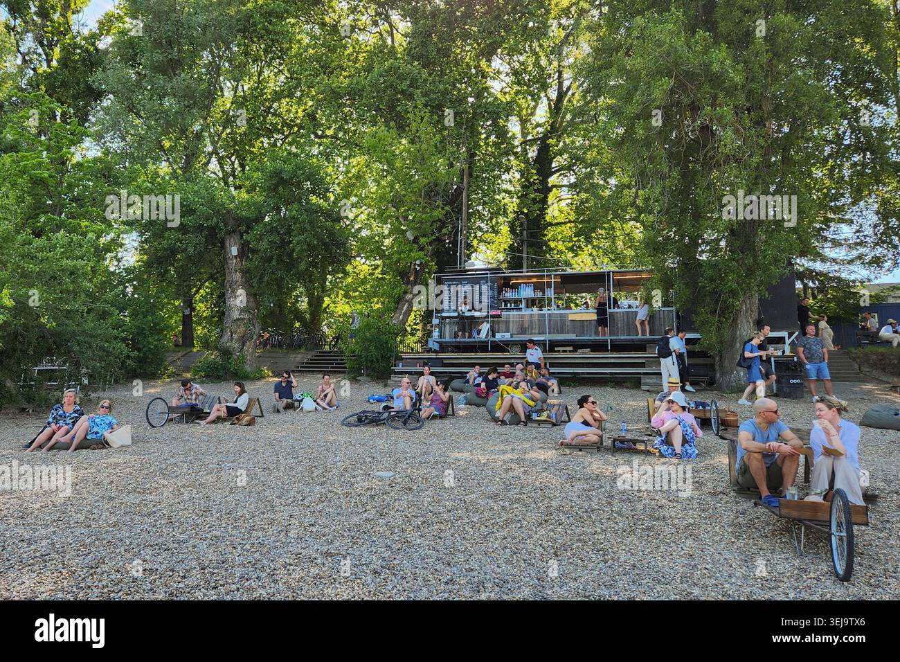 I clienti seduti su una spiaggia di ciottoli di fronte a un bar all'aperto ombreggiato lungo la spiaggia romana di Budapest, Ungheria Foto Stock