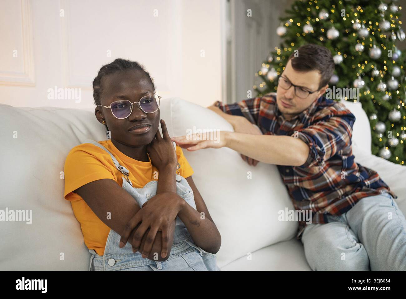Giovane che conforta una triste donna africana su un divano bianco accanto a un albero di natale decorato, indossa una t-shirt arancione e tute in denim, mentre è in un PL Foto Stock