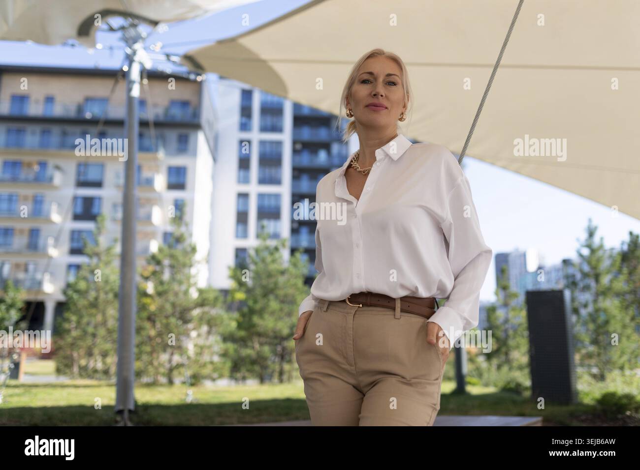 Una donna alla moda si erge tranquillamente all'aperto, indossando una camicia bianca e pantaloni abbronzati, circondata dal verde e da edifici moderni su un luminoso soleggiato da Foto Stock