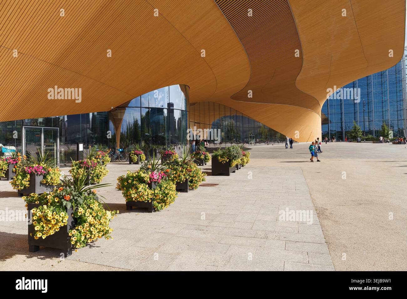 HELSINKI, FINLANDIA - 11 AGOSTO 2025: Baldacchino in legno dorato curvo di un moderno edificio pubblico con piantagioni di fiori in una giornata estiva. Foto Stock