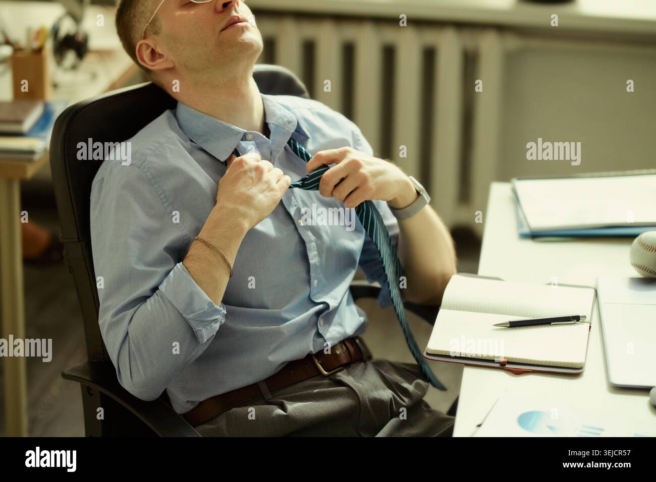 Giovane uomo d'affari caucasico che allenta la cravatta e si appoggia alla scrivania, sentendo lo stress e l'affaticamento del lavoro. Utile per burnout, pressione d'ufficio, lavoro eccessivo, temi di benessere aziendale Foto Stock