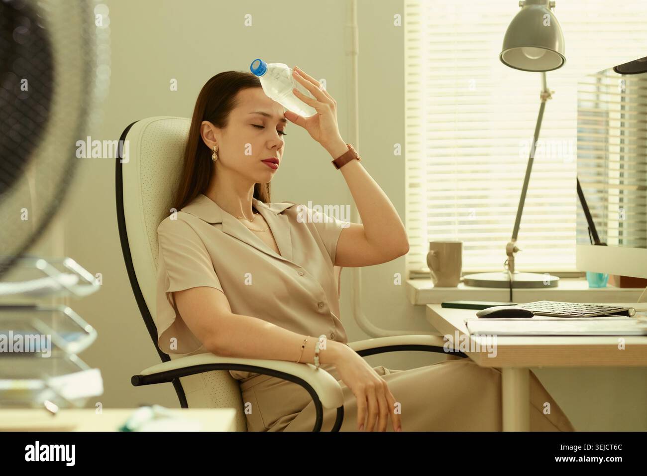 Giovane donna d'affari caucasica che raffredda la fronte con una bottiglia d'acqua seduto alla scrivania dell'ufficio, sperimentando calore stress e stanchezza. Utile per il benessere sul posto di lavoro o per la produttività estiva Foto Stock