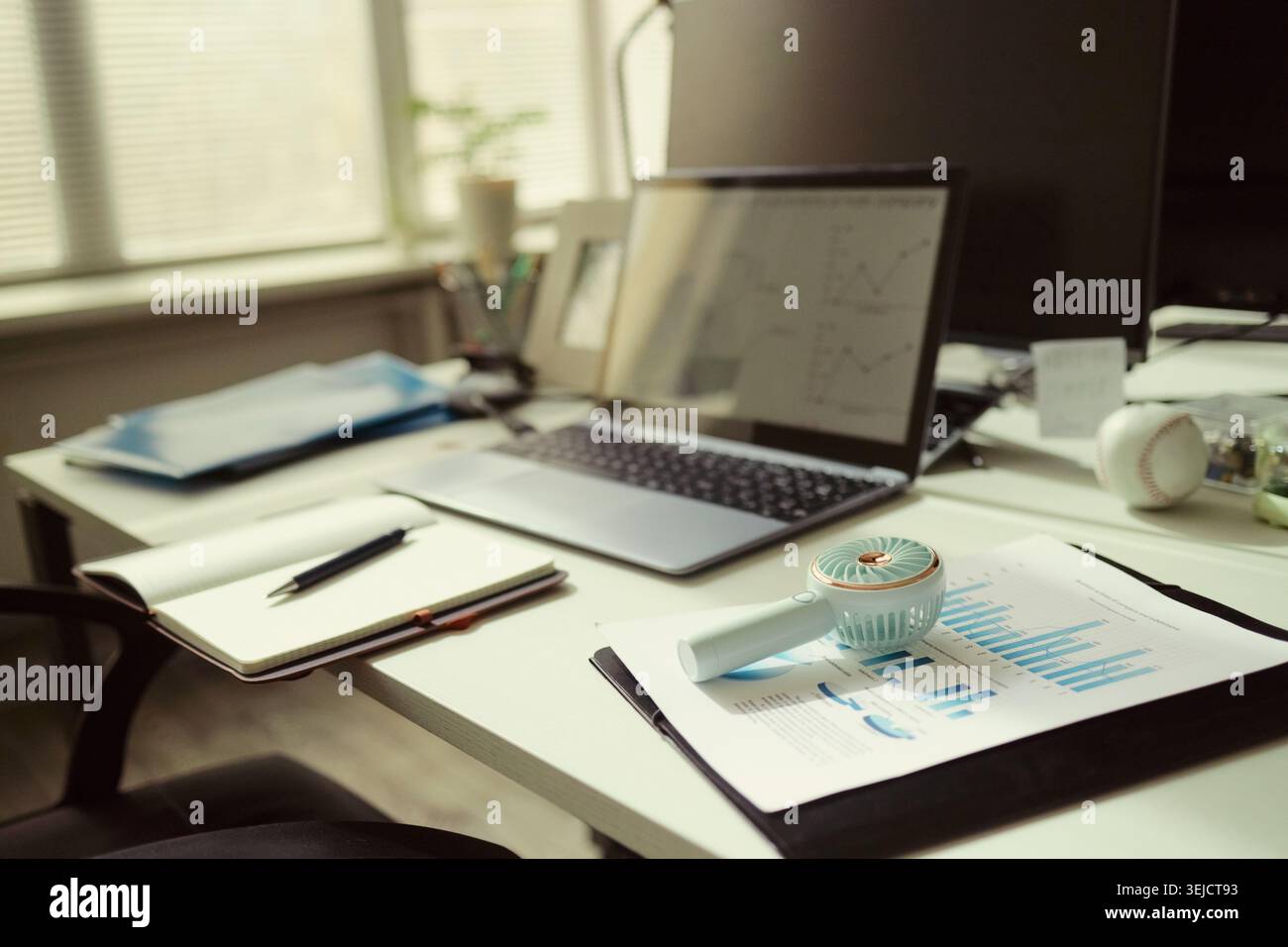 Primo piano della scrivania dell'ufficio con laptop, notebook, penna, grafici, ventola portatile, supporto della pianificazione aziendale e dei contenuti di produttività estiva sul luogo di lavoro per l'uso marketing Foto Stock