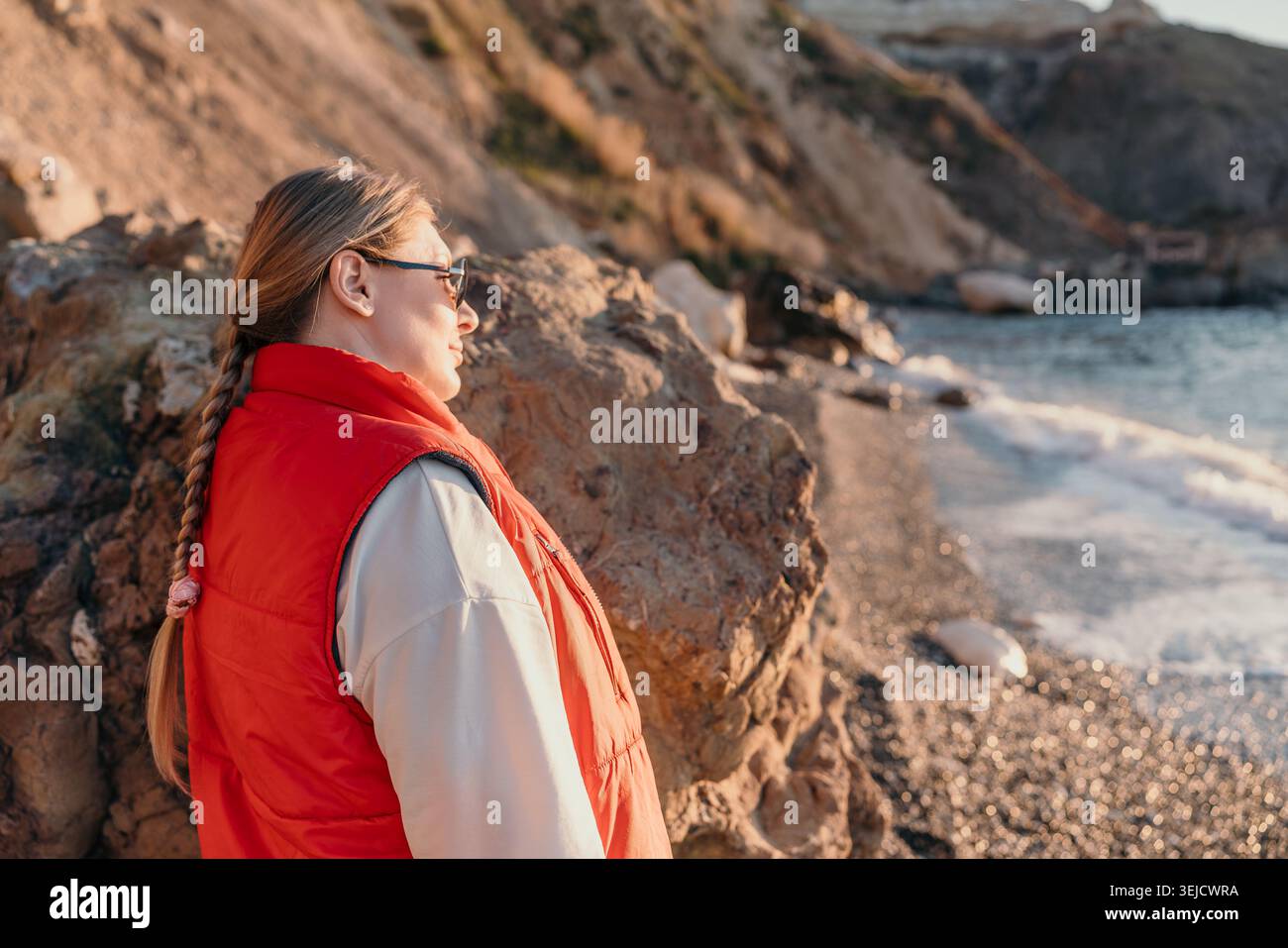 Donna spiaggia costa. Una donna bionda che indossa un gilet rosso guarda le onde dell'oceano da una costa rocciosa durante il tramonto. Foto Stock