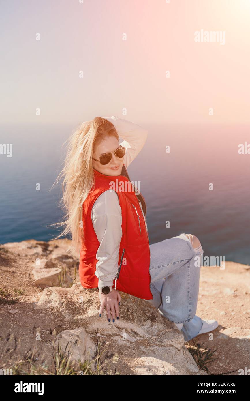 Donna bionda sulla scogliera dell'oceano che indossa un gilet rosso e occhiali da sole seduti su una sporgenza rocciosa che si affaccia sul mare blu al tramonto Foto Stock