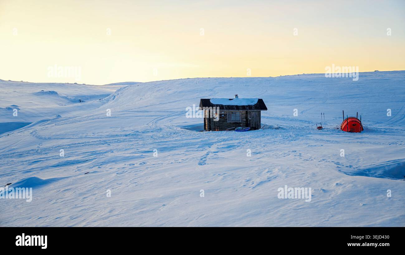 Enontekiö, Finlandia - 09.04.2023: Rifugio in legno isolato e tenda arancione adagiati su un paesaggio bianco e innevato incontaminato durante l'ora d'oro Foto Stock